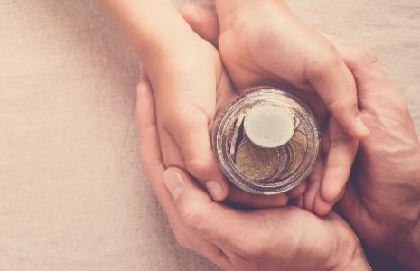 Man's hands cupping a child's hand who is holding a jar with coins inside